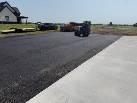 Rural property with newly poured concrete pad, asphalt driveway, farm equipment, and open fields under clear sky