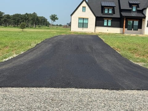 Freshly paved black asphalt driveway leading to a modern two-story house with white and dark exterior on a large grassy lot