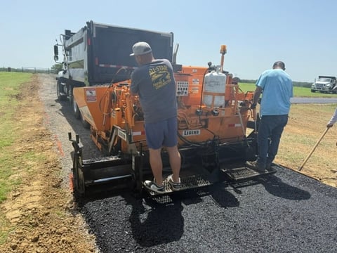 Workers operating an orange asphalt paving machine on a rural road construction site with clear skies