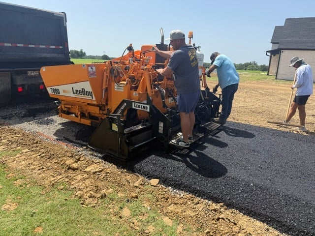 Orange asphalt paving machine laying dark pavement on a rural driveway with workers nearby