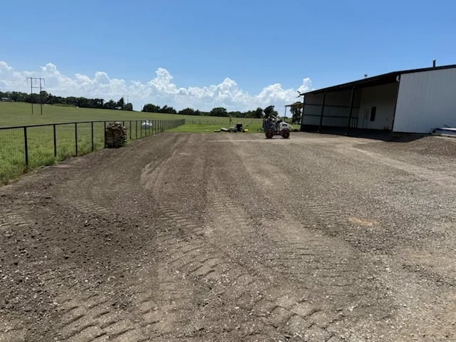 Rural farm driveway with gravel path, fenced pasture on left, metal barn structure on right, and blue sky with white clouds