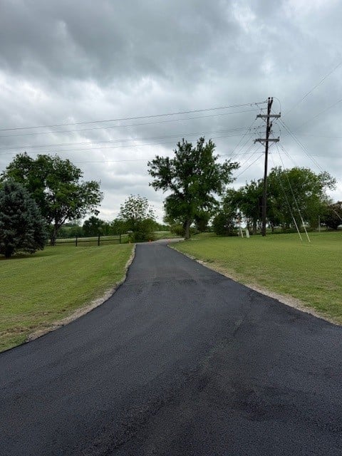 Asphalt driveway curving through a green field with trees and power lines under a cloudy sky
